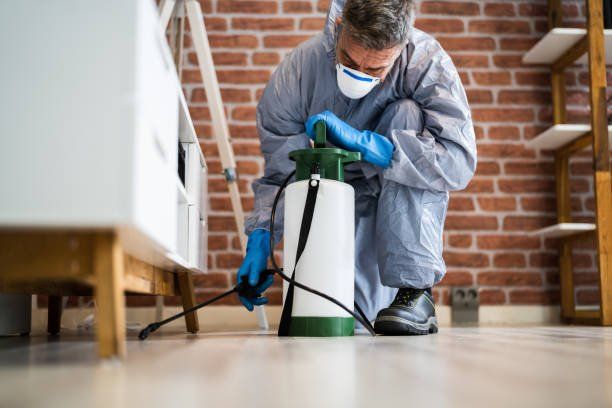 Pest control technician in protective gear spraying along a baseboard indoors